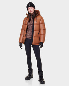 A woman stands against a white background wearing the Rudsak EVIE - L122517 DARK CAMEL down puffer for women with waterproof zippers, black leggings, winter boots, gloves, a patterned top, and a black beanie.