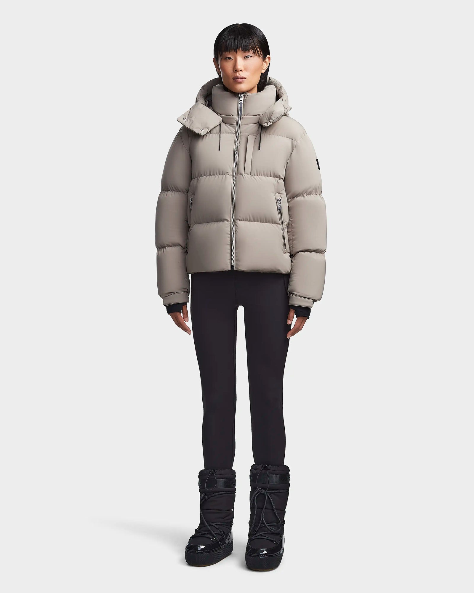 A person stands facing forward against a plain, light background wearing the Rudsak MAVE M - 8123572 Cement womens down puffer jacket, black leggings, and black winter boots.