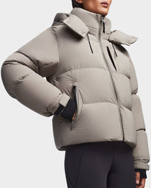 A person stands against a plain background wearing the Rudsak MAVE M - 8123572 Cement womens down puffer jacket with a high collar and zippered pockets, paired with black leggings. The jacket offers warm, lightweight insulation.