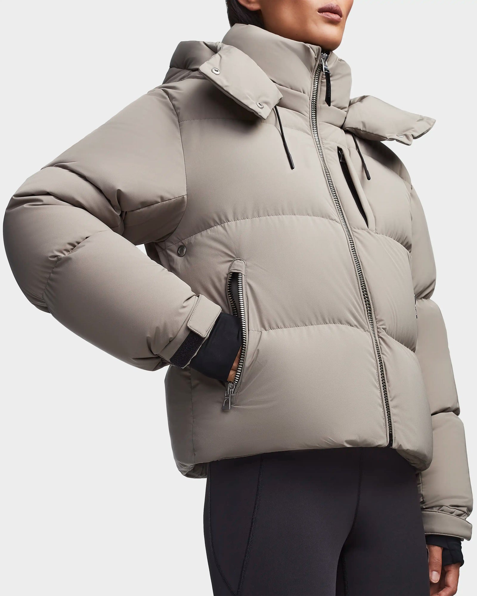 A person stands against a plain background wearing the Rudsak MAVE M - 8123572 Cement womens down puffer jacket with a high collar and zippered pockets, paired with black leggings. The jacket offers warm, lightweight insulation.