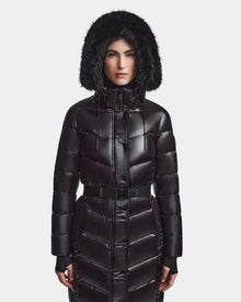 A person wears the Rudsak ELSIE S - L123518 Black women’s maxi down puffer, featuring a shiny finish, belted waist, and a hood with black faux fur trim. They face forward against a plain white background with the hood up.