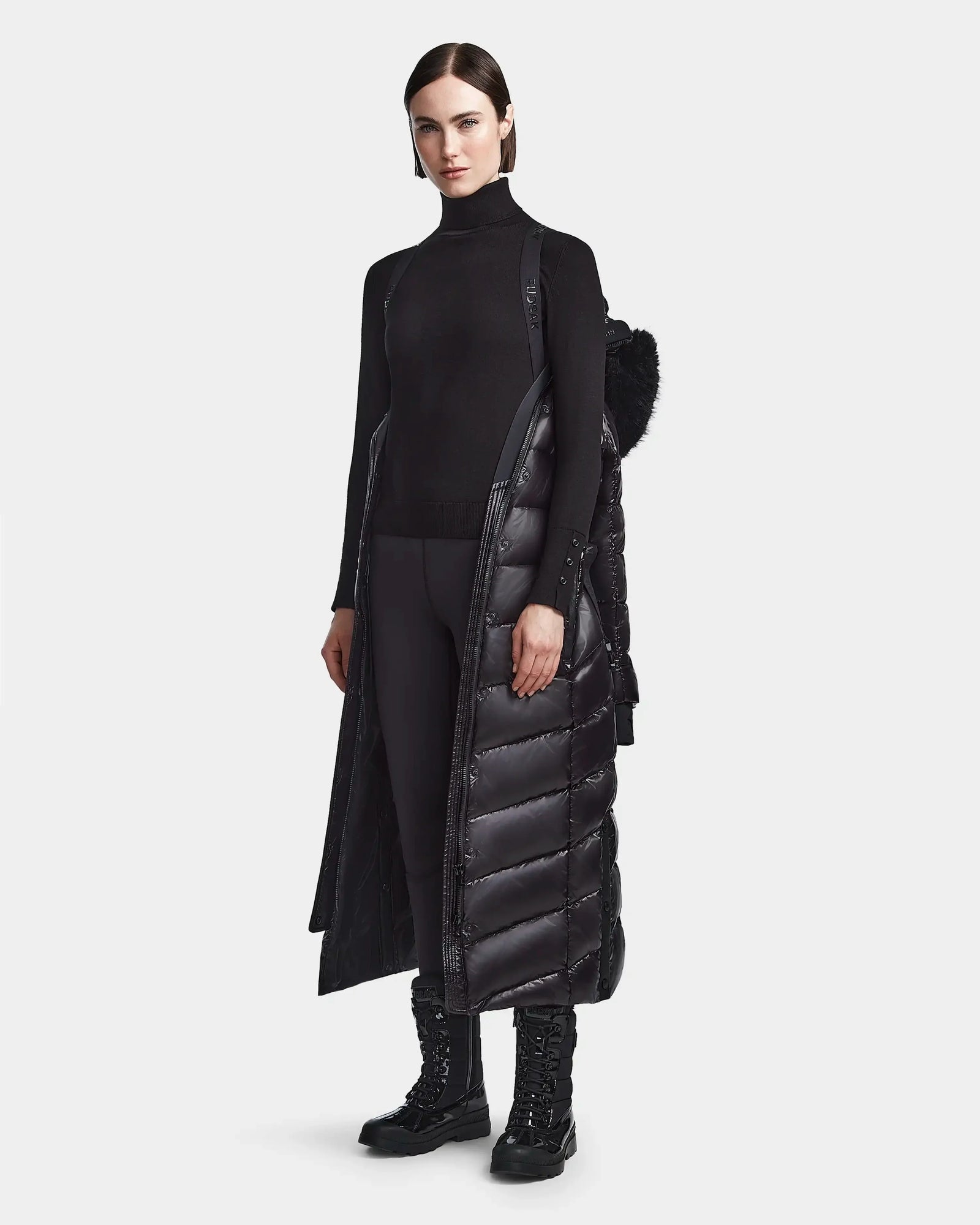 A woman stands against a plain light background wearing the Rudsak ELSIE S - L123518 Black maxi down puffer draped over her shoulders, paired with a black turtleneck, black leggings, and lace-up boots.