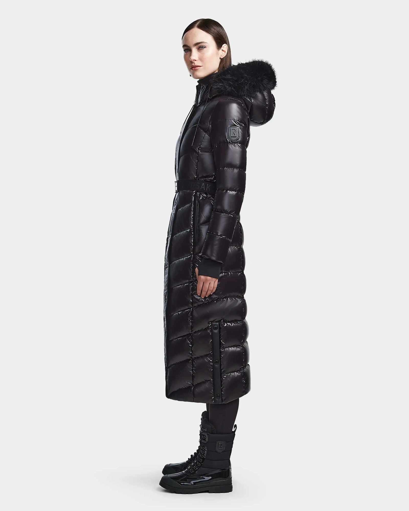A woman stands sideways wearing the Rudsak ELSIE S - L123518 Black maxi down puffer with a fur-trimmed hood, paired with black leggings and lace-up winter boots, against a plain light background.