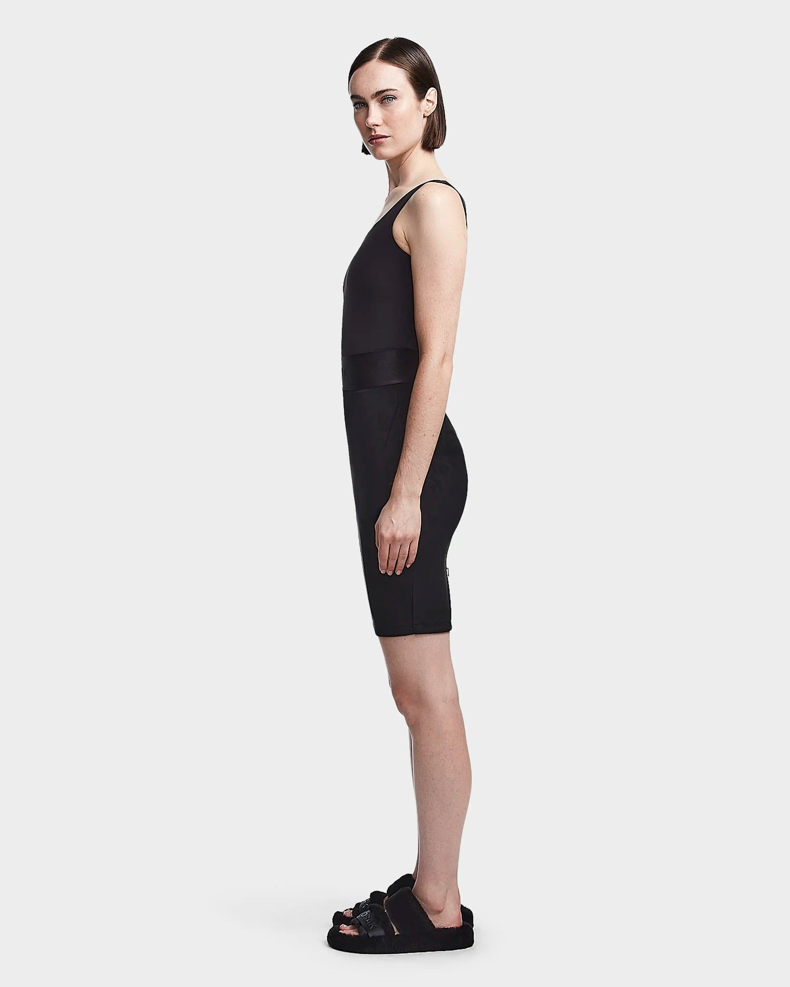 A woman stands in profile against a plain background, dressed in the Rudsak CHEYNA - 8523514 Black sleeveless top and faux suede pencil skirt, paired with matching sandals.