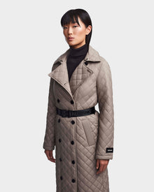 Wearing Rudsak's DAISY - 8123580 Cement women's quilted maxi trench coat with black buttons and a belted waist over a black turtleneck, she stands out against the plain light background in this wind-resistant, water-repellent look.