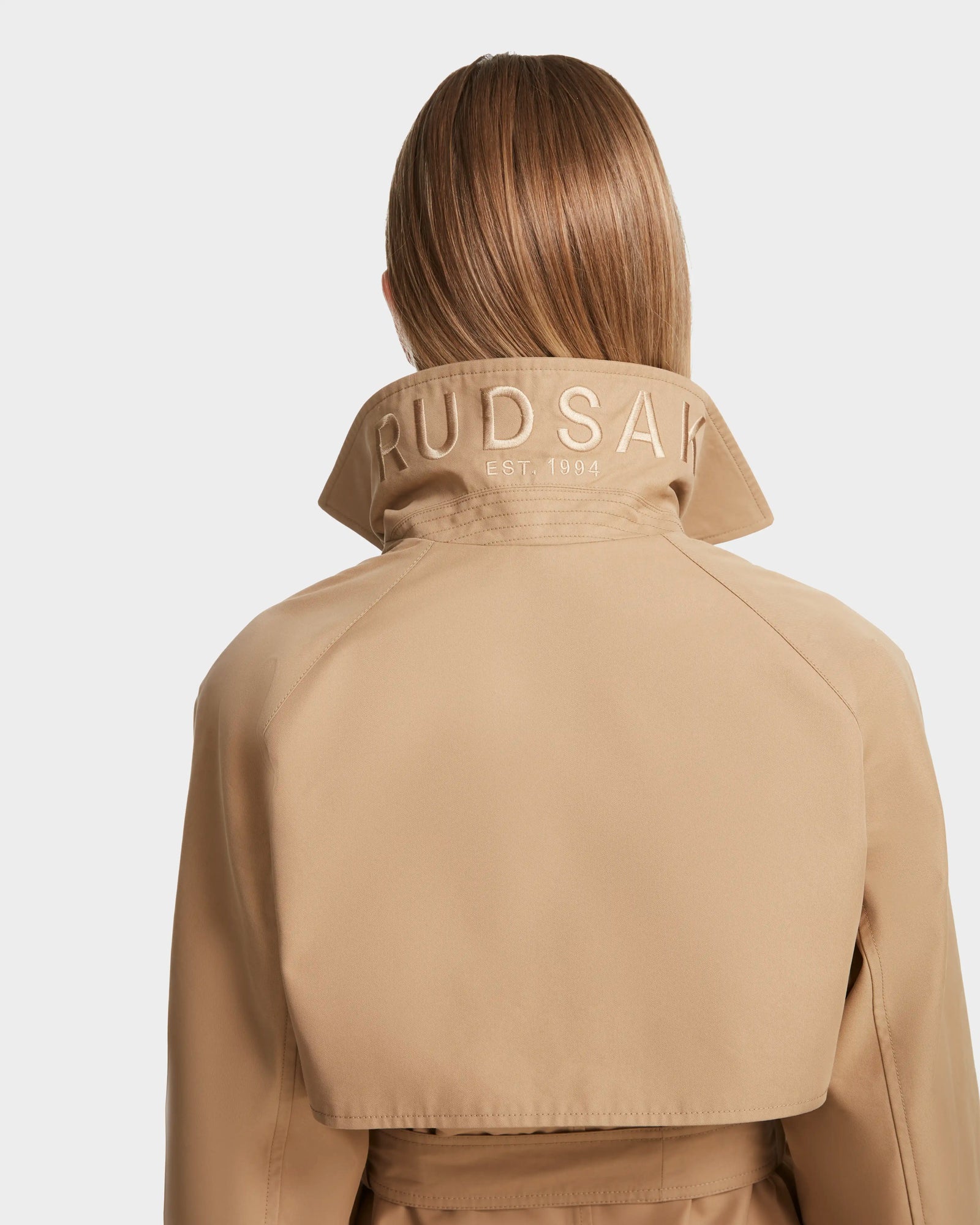 A person with straight, light brown hair is shown from behind wearing the Rudsak MIRELLE - 8124055 Khaki womens trench coat with a tailored silhouette and RUDSAK EST. 1994 embroidered on its turned-up collar.