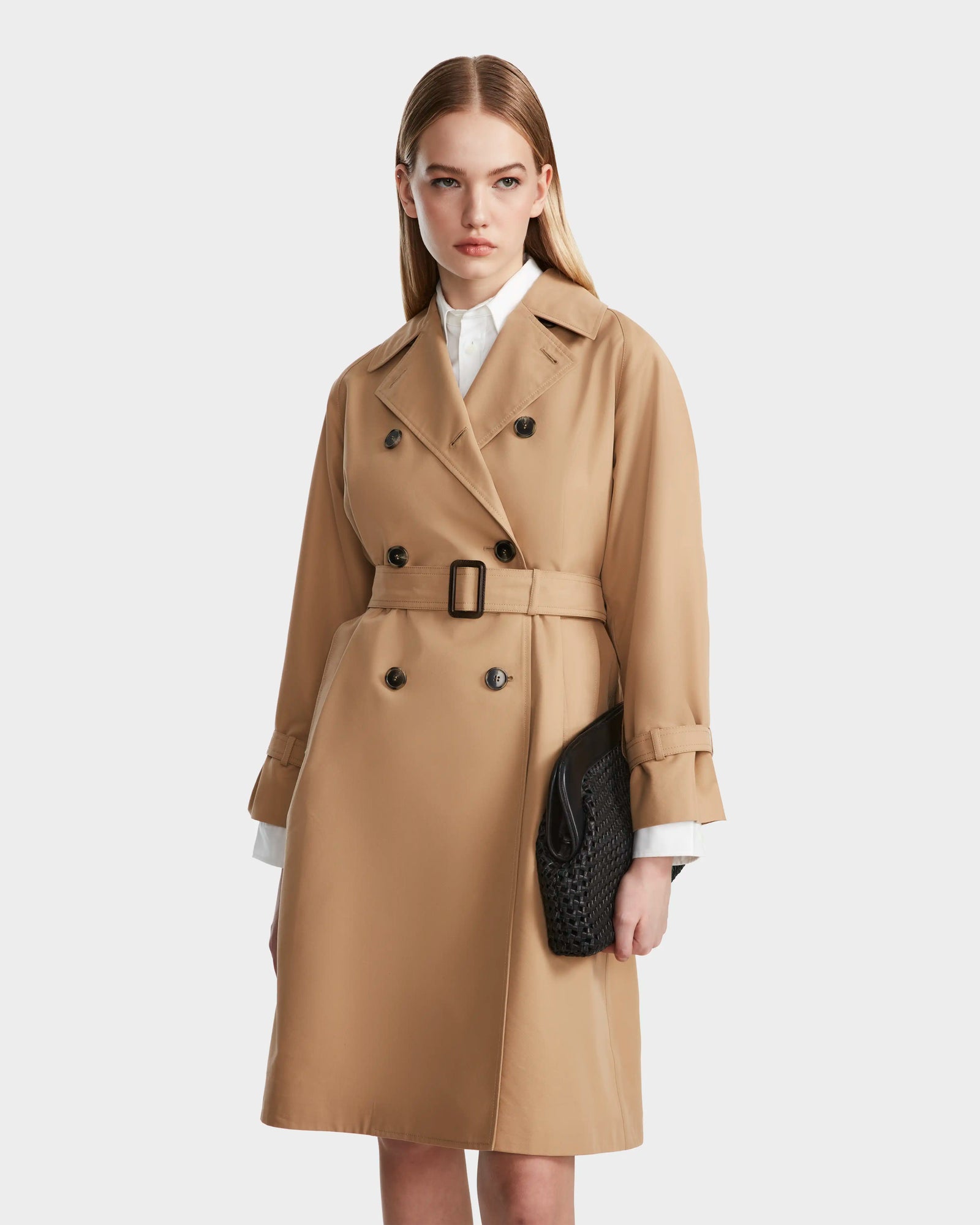 A woman with straight blonde hair models the Rudsak MIRELLE - 8124055 Khaki trench, showcasing its tailored silhouette and double-breasted buttons over a white collared shirt, holding a black woven clutch against a plain light background.
