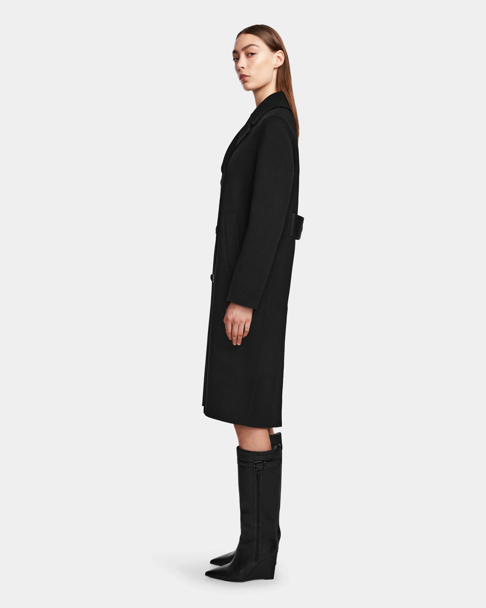 A woman stands in profile against a plain light gray background, wearing the Rudsak LAYLAH - 8124699 Black knee-length tailored wool coat and black knee-high boots with a slight heel.
