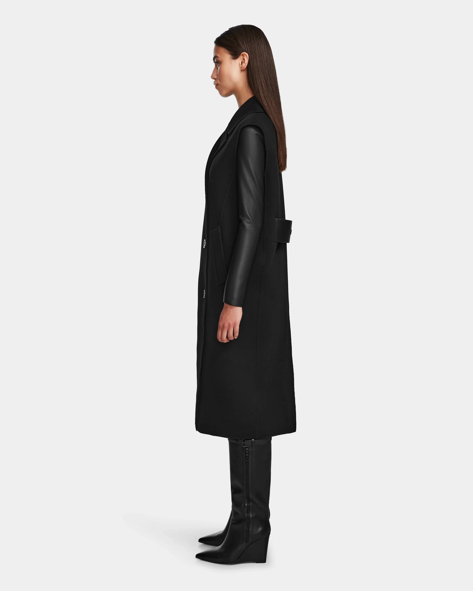 A woman in profile poses against a plain backdrop, wearing the Rudsak NOURA - 8124698 Black knee-length vest over a black leather top, paired with black knee-high boots and sporting straight brown hair.