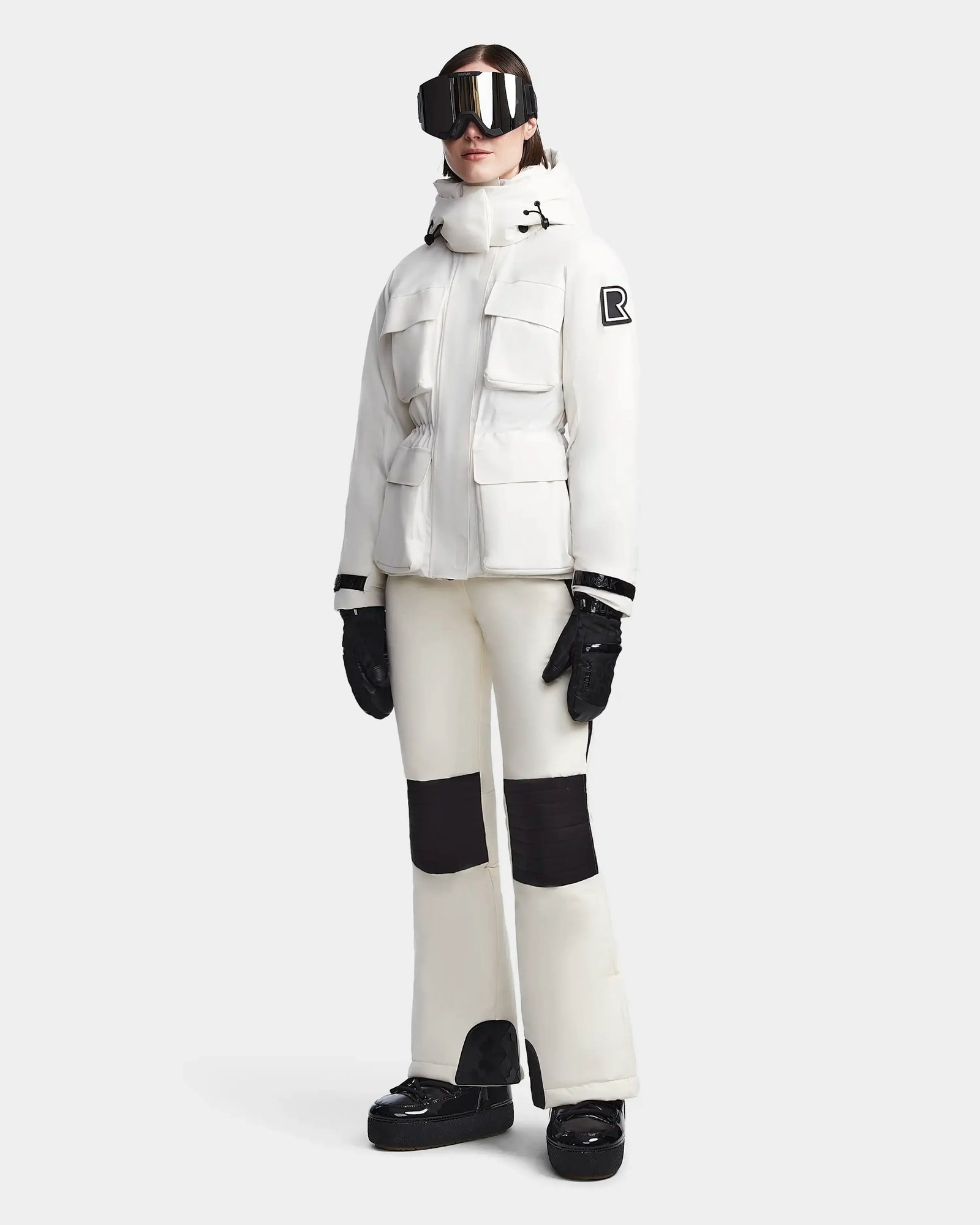 A person wears Rudsak’s LUX - 8123618 Iceland women’s ski jacket with multiple pockets and RECCO, cream ski pants with black knee patches, black gloves, platform boots, and large ski goggles against a plain light background.
