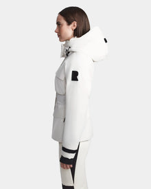 A woman stands in profile wearing the Rudsak LUX - 8123618 Iceland womens ski jacket—a white, hooded winter piece with black accents and matching pants—set against a plain light background.