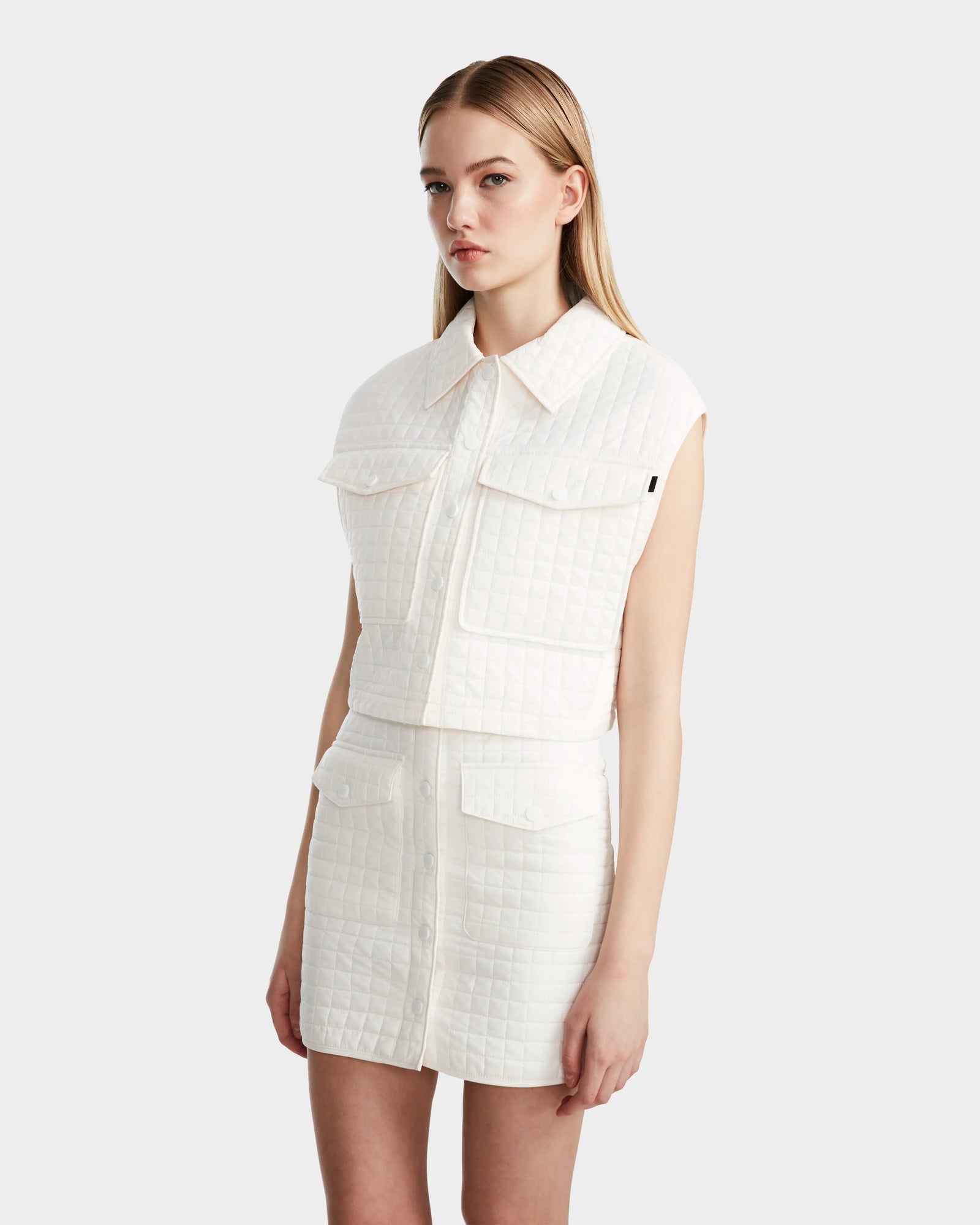 A woman with long blonde hair wears Rudsaks SOLSA - 8124048 Eggshell cropped quilted vest and matching high-waisted skirt, both featuring eco-friendly Sorona fill, while standing against a plain light background.