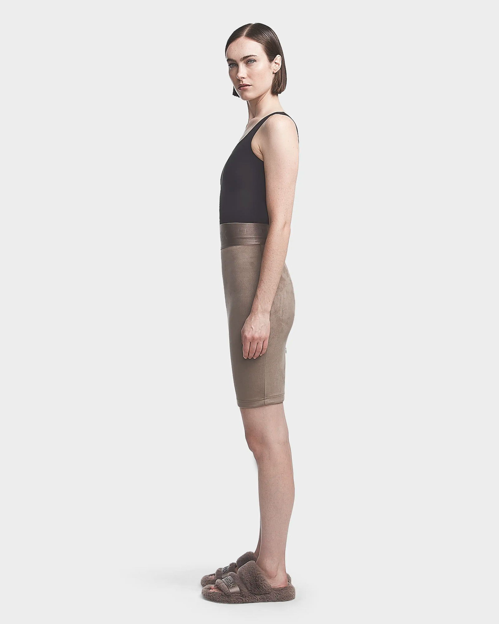 A woman stands in profile against a plain background, wearing the Rudsak CHEYNA - 8523514 Cement skirt with a fitted silhouette and elasticized waistband, paired with a black sleeveless top and brown fuzzy slide sandals.