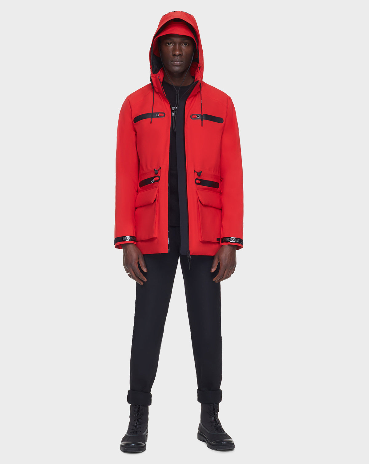 A person stands facing forward wearing the RUDSAK JACY - 6122000 POPPY RED raincoat with black zippers and pockets, a matching red cap, black pants, and black boots against a plain white background.