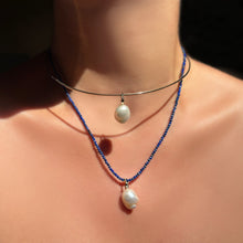 A layered look of a mini baroque pearl charm on a gold wire choker over a lapis slim gemstone necklace with a 2nd mini baroque pearl, shown on the neck in sunlight.