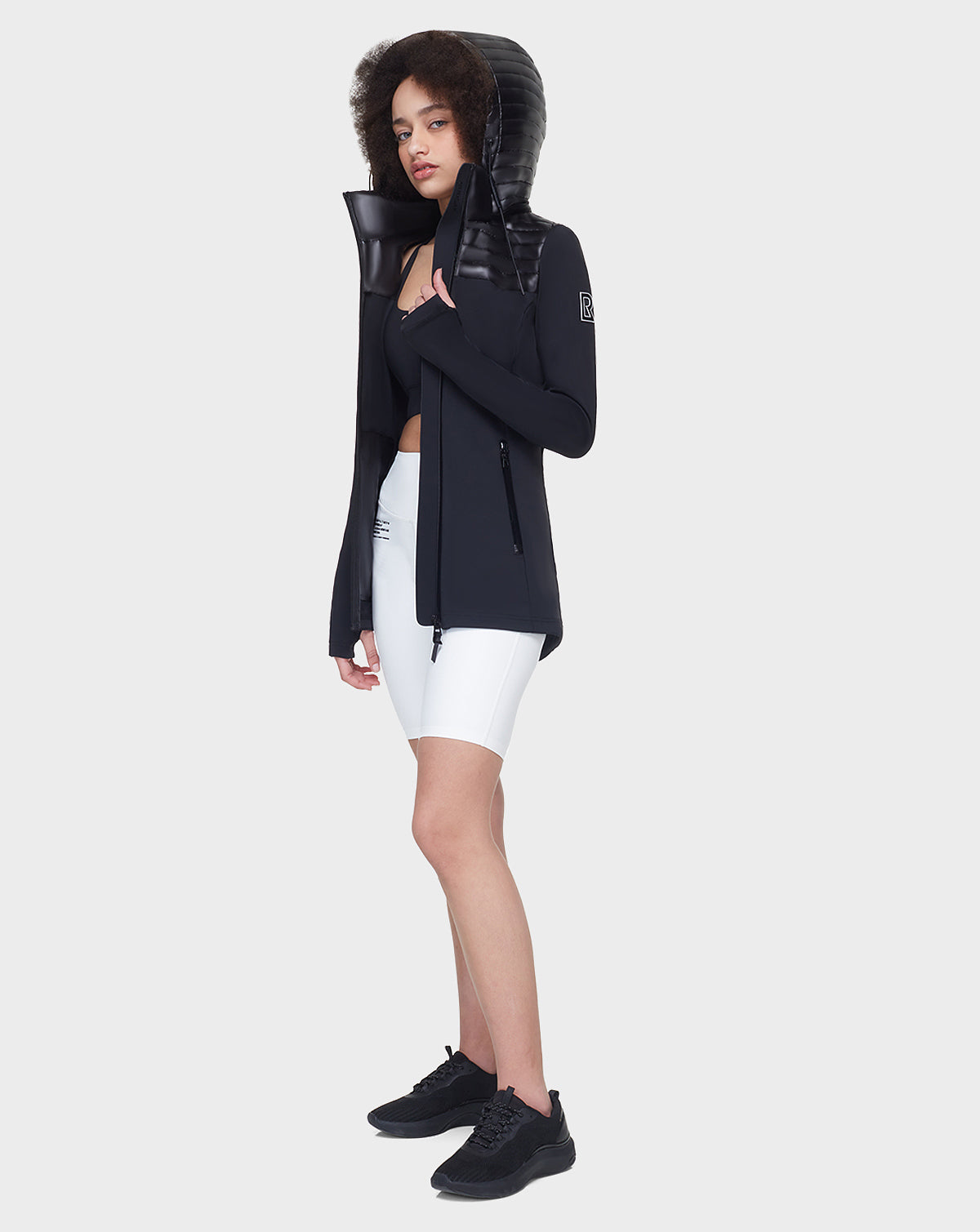 A woman with curly hair wears the Rudsak CAMIRA - 8122011 BLACK activewear jacket with waterproof zippers over a black sports bra, white biker shorts, and black sneakers, standing against a white background and looking ahead neutrally.