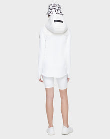 A person stands facing away against a plain light gray background, wearing the CAMIRA - 8122011 WHITE jacket by Rudsak, coordinated with white cycling shorts, white sneakers, and a black-and-white patterned bucket hat—ready for outdoor workouts.