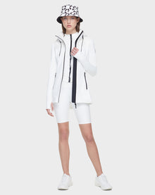 A person stands facing forward in Rudsaks CAMIRA - 8122011 WHITE womens activewear jacket, paired with white cycling shorts, sneakers, and a white bucket hat with a black geometric pattern—perfect for outdoor workouts on a light gray background.